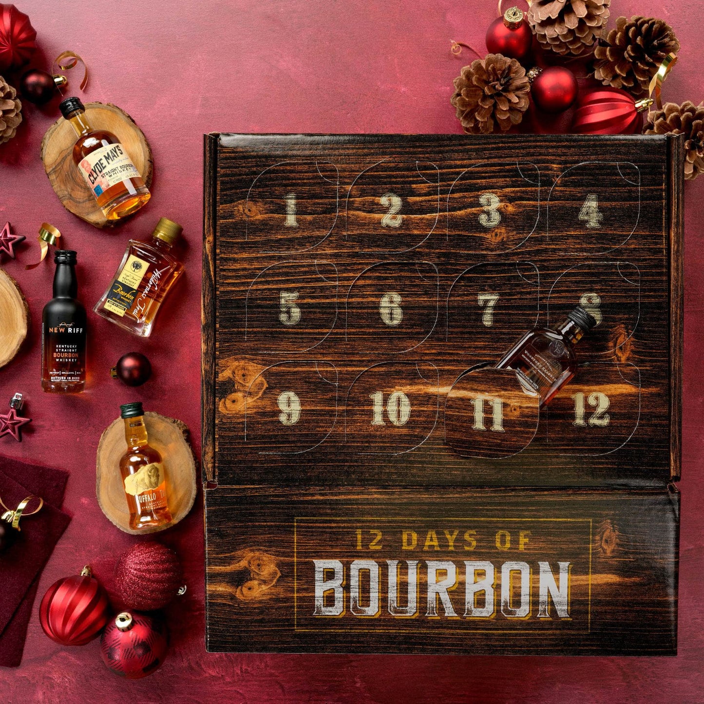 A 12-day bourbon advent calendar styled on a textured red surface with holiday decorations and miniature bourbon bottles around it. The calendar is dark brown wood with numbered circular compartments and the text '12 DAYS OF BOURBON' on the front.