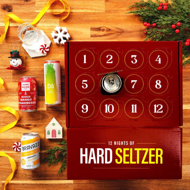 A Christmas-themed hard seltzer advent calendar on a wooden table, surrounded by festive props like pine branches, candy canes, a snowman ornament, and yellow ribbons. The red calendar box shows numbered compartments with a seltzer can inserted in the center, and six sample seltzer cans are lined up to the left.