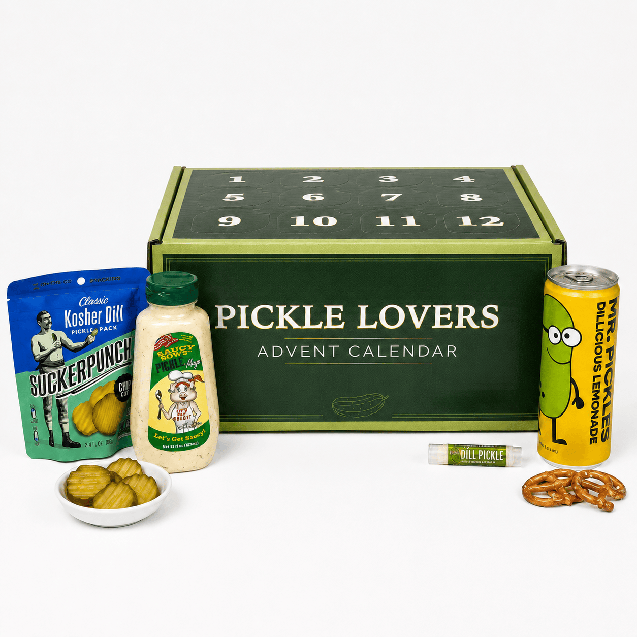 Pickle Lovers Advent Calendar gift set on a white background, featuring a dark green numbered box with mustard-green trim, surrounded by themed snacks including a bag of Kosher Dill pickles, a jar of relish, a can of lemonade, pretzels, and a small bowl of pickled onions.