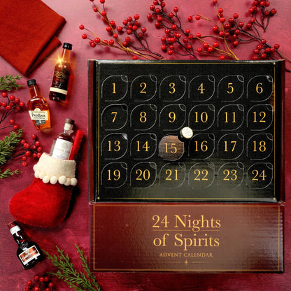 A festive flat-lay of a '24 Nights of Spirits' advent calendar on a dark red background, surrounded by holiday decorations including red berries, pine branches, a red stocking, and miniature liquor bottles.