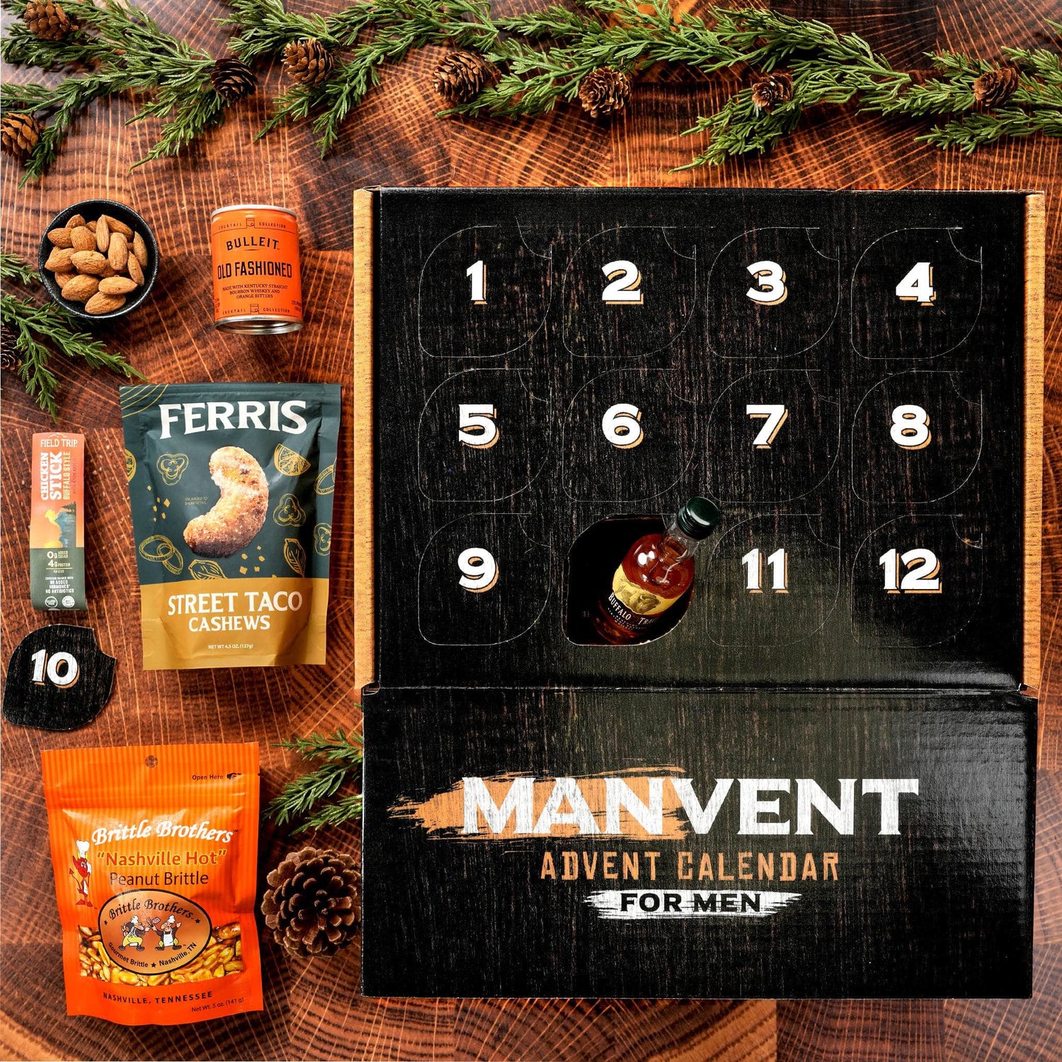 A Manvent Advent Calendar for Men displayed on a rustic wood background with pine branches and pinecones. The black numbered calendar box is open, showing several snack compartments, with some snacks like Ferris Street Taco Cashews and Brittle Brothers Peanut Butter already opened and arranged beside it.