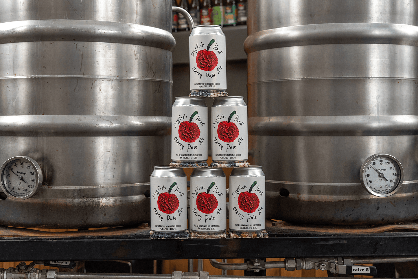 Stack of Dogfish Head Cherry Pale Ale beer cans in front of metal beer tanks