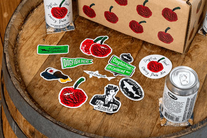Dogfish Head Stickers and a can on a wooden barrel with a box.