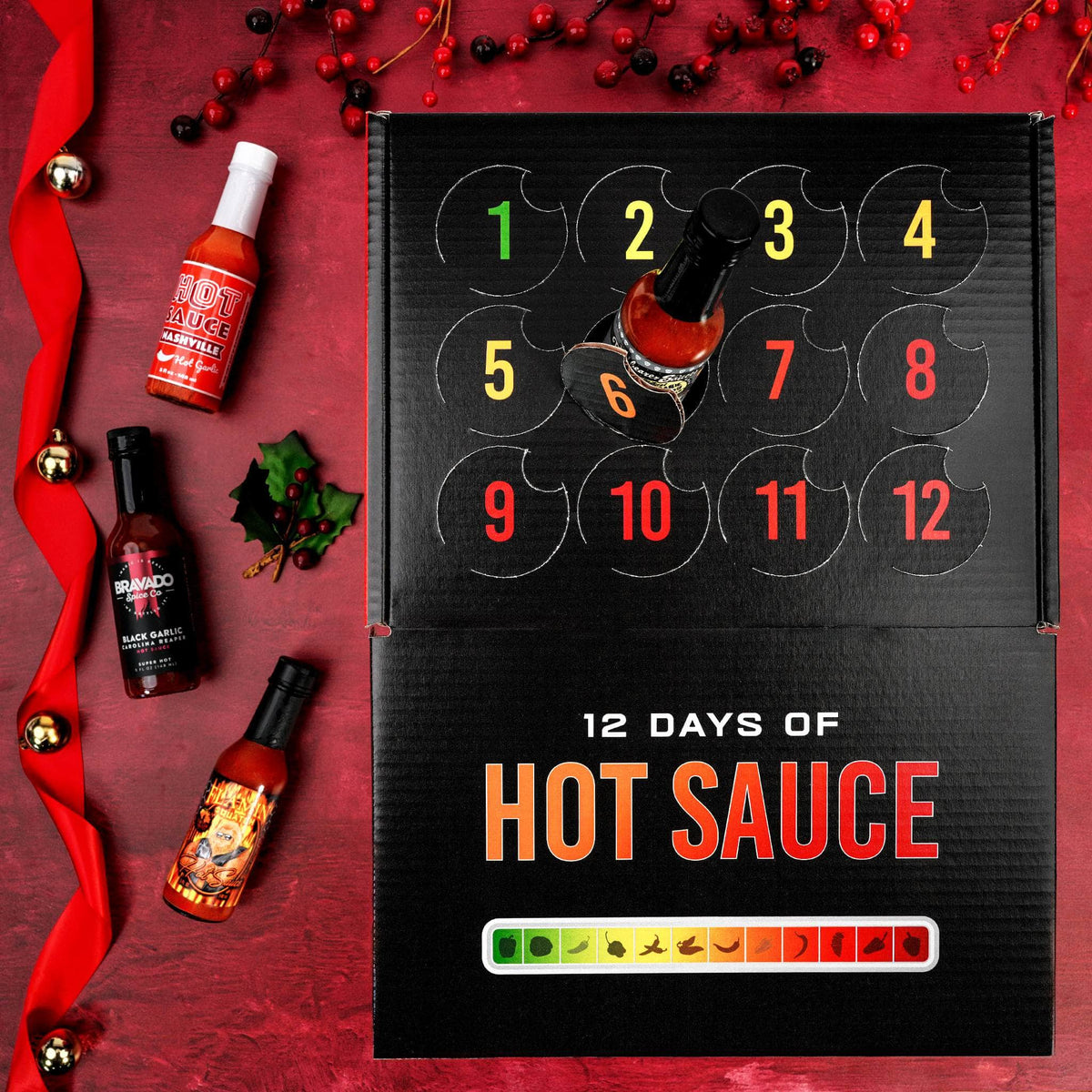 A 12-day hot sauce advent calendar box on a red holiday-themed background, surrounded by individual hot sauce bottles, red berries, and a red ribbon.