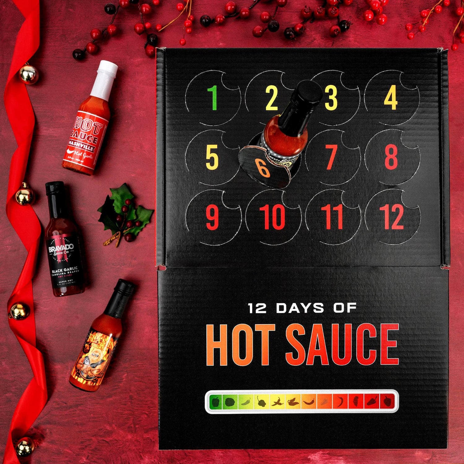A 12-day hot sauce advent calendar box on a red holiday-themed background, surrounded by individual hot sauce bottles, red berries, and a red ribbon.
