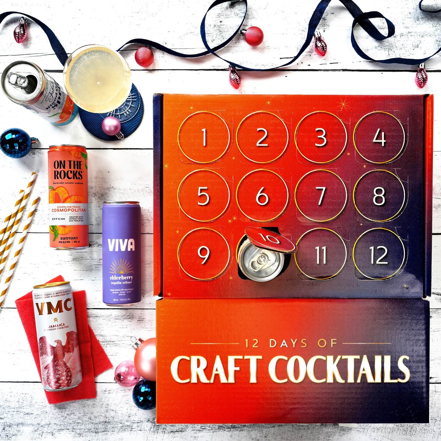 An advent-style cocktail gift box labeled '12 DAYS OF CRAFT COCKTAILS' sits on a white wood surface with several cocktail cans and drink accessories arranged around it. The main product is a red-to-purple numbered box with 12 circular compartments, and one compartment is open, showing a can inside.