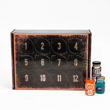 A 12-day moonshine advent calendar box with numbered compartments on the front, shown on a white background with three small jars of colorful spices or candies beside it.