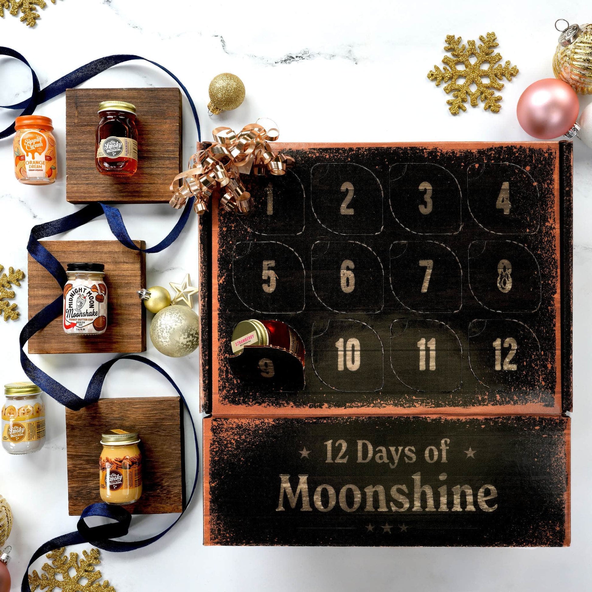 A Christmas advent calendar labeled '12 Days of Moonshine' in a rustic black and dark wood finish, displayed on a white marble surface with decorative gold ornaments, a gold snowflake, a navy ribbon, and several small jars of moonshine arranged around it.