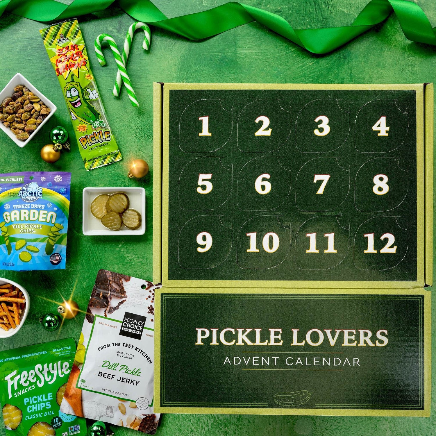 Pickle Lovers Advent Calendar shown open on a green surface with holiday snacks and candy props around it. The calendar has 12 numbered compartments on the top panel and the words 'PICKLE LOVERS ADVENT CALENDAR' on the bottom panel.