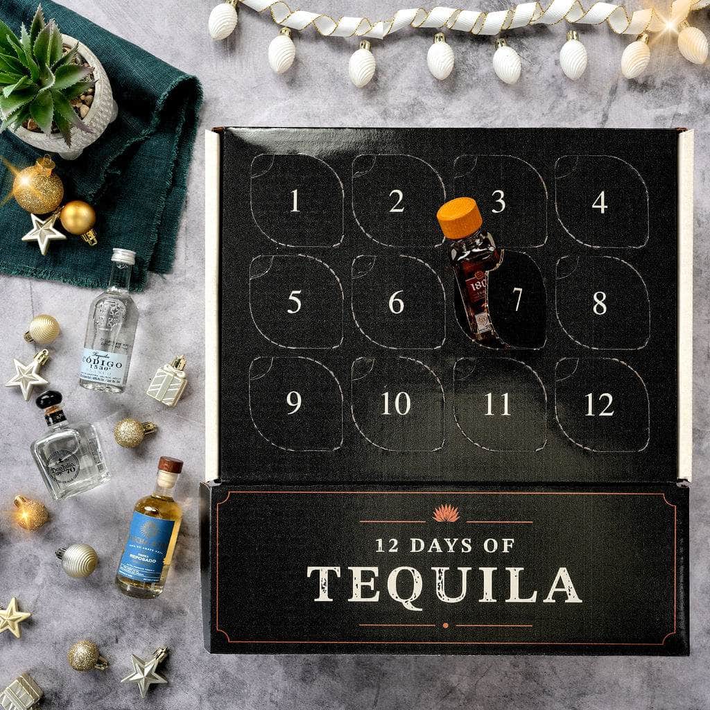 A 12-day tequila advent calendar box shown open on a gray stone surface, with 12 numbered compartments and one small tequila bottle placed in the center compartment. The box is decorated with festive props including miniature bottles, ornaments, a small plant, and string lights.