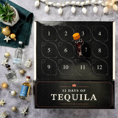 A 12-day tequila advent calendar box shown open on a gray stone surface, with 12 numbered compartments and one small tequila bottle placed in the center compartment. The box is decorated with festive props including miniature bottles, ornaments, a small plant, and string lights.