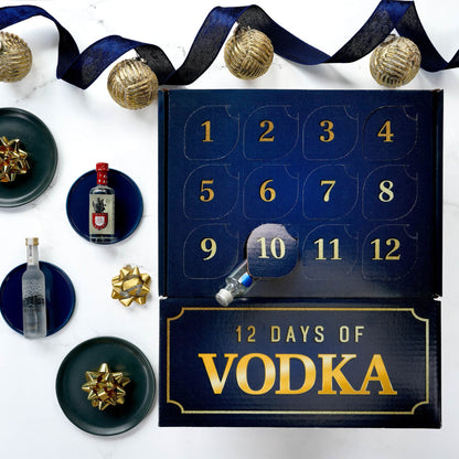 A 12-day vodka advent calendar laid flat on a white marble surface, surrounded by decorative navy ribbon, gold ornaments, and small plates with bottles and gift bows. The calendar is dark blue with gold numbering and the text '12 DAYS OF VODKA' on the front.