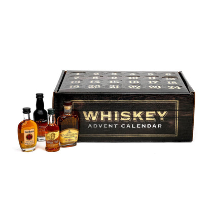 A whiskey advent calendar featuring a large black wooden box with gold lettering that reads 'WHISKEY ADVENT CALENDAR' and twenty numbered compartments on top, shown with four miniature liquor bottles in front on a white background.
